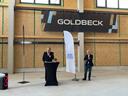 Zwei Männer halten eine Rede vor dem Schild "Goldbeck"