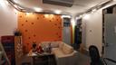 Ein kleines Büro, links eine weiße Wand, einige Getränkekasten und eine Zimmerpflanze. Mittig im Bild eine orange Wand, darauf rote Blumenmuster. Rechts einige Regale, eine graue Tür, ein Bürostuhl. Vorne in der Bildmitte eine Couchecke, auf dem Couchtisch Pinsel und Farben. Darüber eine Lichterkette, die in warmen Farben leuchtet. Am Bildrand eine Malerleiter.