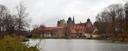 Schlossanlage aus hellem Stein mit mehreren Rundtürmen und roten Dächern im Markt Thurnau, direkt an einem ruhigen Gewässer, dem Thurnauer Schlossweiher, gelegen. Bäume mit dünnem Herbstlaub und kleinere Gebäude umgeben die Anlage. Der Himmel ist grau und bedeckt.
