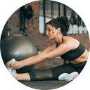 A woman is sitting on the floor in a gym and stretching.