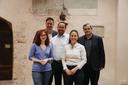 Fünf Erwachsene stehen in einem Raum mit einer hellen Wand. Oberhalb der Gruppe sind Spuren von historischen Wandmalereien. Links ist eine Holztür zu sehen. Die Personen stehen in zwei Reihen, zwei Frauen vorne und drei Männer dahinter, und schauen zur Kamera.