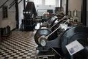 Historischer Maschinenraum mit großen metallenen Motoren und Kompressoren in einer Reihe, einem großen Schwungrad mit Druckanzeigen und schwarz‑weiß gefliestem Boden.