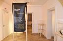 Ein heller Ausstellungsraum zeigt ein großes schwarzes Banner, das in deutscher und arabischer Sprache beschreibt, wie Krieg unerwartet in den Alltag einbricht und Menschen unvorbereitet trifft. Das Banner steht in einem Holzrahmen, dahinter befinden sich leere Glas‑Holz‑Vitrinen. Links im Raum ist eine weiße Tür mit der Beschriftung ‚BMZ‘ zu sehen. Die Beleuchtung ist warm und richtet sich auf die Ausstellungselemente.