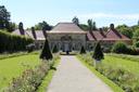 Schlossanlage aus hellem Sandstein mit roten Dächern und zahlreichen Fenstern. Ein breiter Kiesweg führt durch einen blühenden Garten direkt zum Tor, das den Eingang zum Festsaal darstellt..