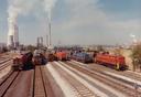 Mehrere Diesellokomotiven stehen nebeneinander auf parallelen Gleisen in einem Bahnbetriebsbereich. Im Hintergrund sind Industriegebäude mit hohen Schornsteinen zu sehen, aus denen heller Rauch aufsteigt. Die Umgebung besteht aus offenen Feldern unter einem klaren Himmel.