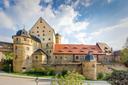 Ansicht von Schloss Thurnau von der Straßenseite her. Das Schloss ist gebaut aus hellem Stein. Mehrere Rundtürme mit spitzen Dächern. Ein höheres Hauptgebäude mit kleinen Fenstern. Teile des Dachs sind rot gedeckt. Die Burg ist von einer Steinmauer und Grünflächen umgeben. Im Vordergrund ein gepflasterter Weg und Laternen. Der Himmel ist leicht bewölkt und die Szene ist sonnig beleuchtet.