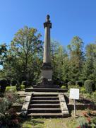 Die Ottosäule in Ottobrunn, ein hohes, geriffeltes Steindenkmal mit einer Büste König Ottos von Griechenland an der Spitze. Am Sockel befinden sich steinerne Löwenfiguren und eine Inschrift. Eine Treppe führt zum Denkmal hinauf, das von Bäumen, gepflegten Sträuchern und Blumen umgeben ist. Darüber erstreckt sich ein wolkenloser, blauer Himmel.