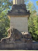 Detailaufnahme des Sockels der Ottosäule in Ottobrunn. Ein steinerner Löwe liegt vor einer Gedenkinschrift, die an die Abschiedsstunde König Ottos I. von Griechenland und seines Vaters Ludwig I., König von Bayern, erinnert. Über der Inschrift befinden sich kunstvolle steinerne Verzierungen. Im Hintergrund sind Bäume zu sehen.