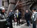 In einer weitläufigen Industriehalle stehen mehrere Personen zwischen großen Metallrohren und technischen Anlagen. Eine Person trägt Kopfhörer und hält eine Tonangel, als Teil einer Film‑ oder Dokumentaraufnahme. Die übrigen Personen tragen Winterjacken, Schals und Mützen und beobachten die Szene. Die Umgebung wirkt kalt und funktional, mit sichtbaren Leitungen und Maschinen im Hintergrund.