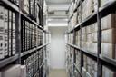 Schmaler Archivgang mit Metallregalen: links dicht gefüllte Ordnerreihen, rechts gestapelte beschriftete Archivkartons.
