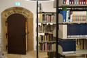 Bibliotheksraum mit Metallregalen voller Bücher rechts und einer braunen Tür in einem steinernen Rahmen links. Über der Tür steht eine Inschrift und ein grünes Notausgangsschild.