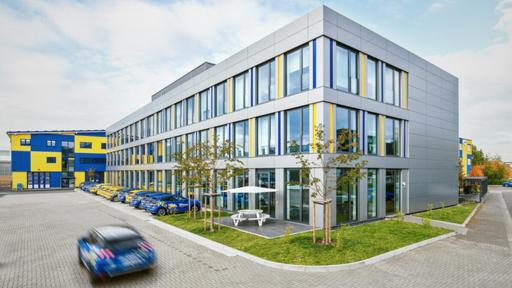 Das neuste Bürogebäude der MAINGAU Energie GmbH mit einer grauen Fassade und großen Fenstern, die mit gelben und blauen Akzenten verziert sind. Im Vordergrund befindet sich eine kleine Terrasse mit einem weißen Sonnenschirm und einer Sitzgelegenheit. Auf dem Parkplatz stehen mehrere Fahrzeuge mit auffälligen, gelb-blauen Designs. Links im Bild ist ein weiteres Gebäude mit einer gelb-blauen Fassade sichtbar.