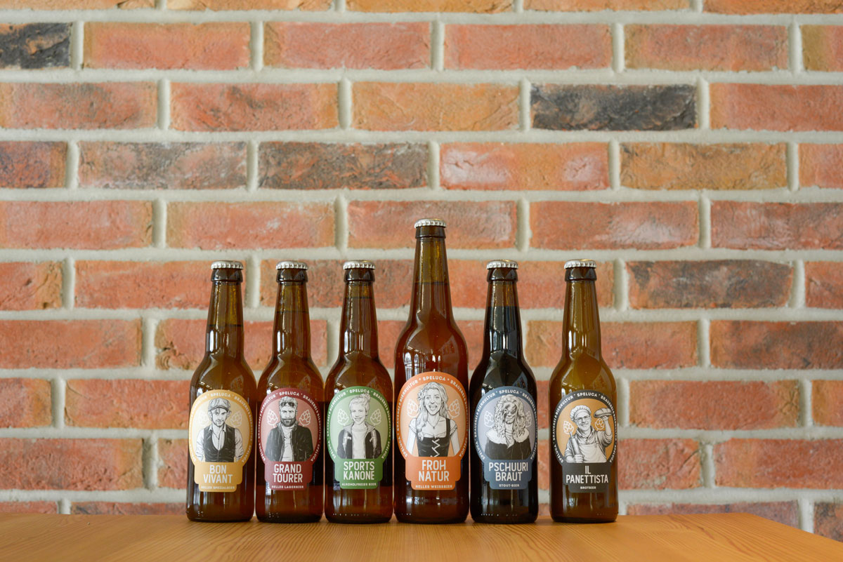 Verschiedene Bierflaschen der Speluca Brauerei stehen vor einer Wand aus rotem Sichtmauerwerk.