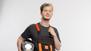 Ein Mann steht vor einer weißen Wand und trägt eine schwarz-orange Latzhose