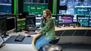 Eine junge Frau sitzt an einem Schreibtisch mit vielen Monitoren und telefoniert. 