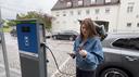 Astrid Drechsel-Grau schaut auf die Ladekarte und hält ihr Handy in der Hand. Im Hintergrund ist das Elektroauto zu sehen, welches nun mithilfe des Ladekabels mit der Ladesäule verbunden ist.