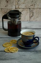 Es ist eine Tasse Tee mit einer Zitronenscheibe zu erkennen. Im Hintergrund liegen weitere Zitronenscheiben und eine durchsichtige Teekanne.