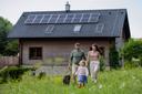 Eine Familie mit Hund geht lächelnd über eine Wiese. Im Hintergrund ist ein Haus mit Photovoltaikanlage auf dem Dach.