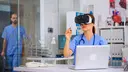 Eine Ärztin in blauer Krankenhauskleidung trägt eine Virtual-Reality-Brille und sitzt an einem Schreibtisch mit Laptop. Im Hintergrund ist ein weiterer medizinischer Mitarbeiter in einem modernen Klinikraum zu sehen.