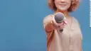 A woman holds a microphone directly towards the camera, with a blurred blue background.