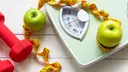 A scale with apples on it and a measuring tape and dumbbells next to it for a weight loss concept