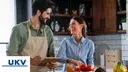 A man and a woman are standing in the kitchen and are amiling at eachother, in front of them stands a juice, tomates and bananas with clientlogo UKV