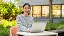 Person sitting at an outdoor table working on a laptop, with greenery and a building in the background.