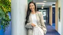 Person leaning against a concrete pillar in a modern office hallway, wearing a light blazer and white trousers.