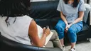 Woman sits on the sofa and another woman sits opposite her in an armchair, holding a notepad and a pen for taking notes.