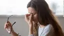 Woman puts her left hand between her eyes, her face looks painful, in the right hand she is holding glasses