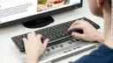 Person von hinten, die am Computer sitzt und eine Tastatur und eine Braillezeile benutzt.