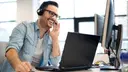 Laughing young man at work on a laptop, talking on the phone via a headset