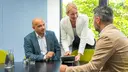 Three people sitting and standing at a meeting table, with one person showing something on a tablet to the others.