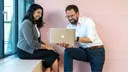 Two people sitting in front of a pink wall, looking together at an open laptop.