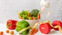 Vegetables, a measuring tape, a water bottle with water and a 2 kg dumbbell on a white table
