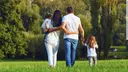 A familiy with four family members ist walking in the green nature, you see them from behind. The mother is holding a little boy in her arms and the father is holding a little gril at his right hand. 