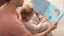 A man is sitting on the floor with a book for children in his hands and in front of him is sitting a little girl