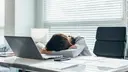 Tired, young woman in the office at her workplace, resting her head on the table in front of her laptop