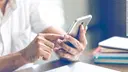 A man is sitting at a table, holding a smartphone in his hands and watching it