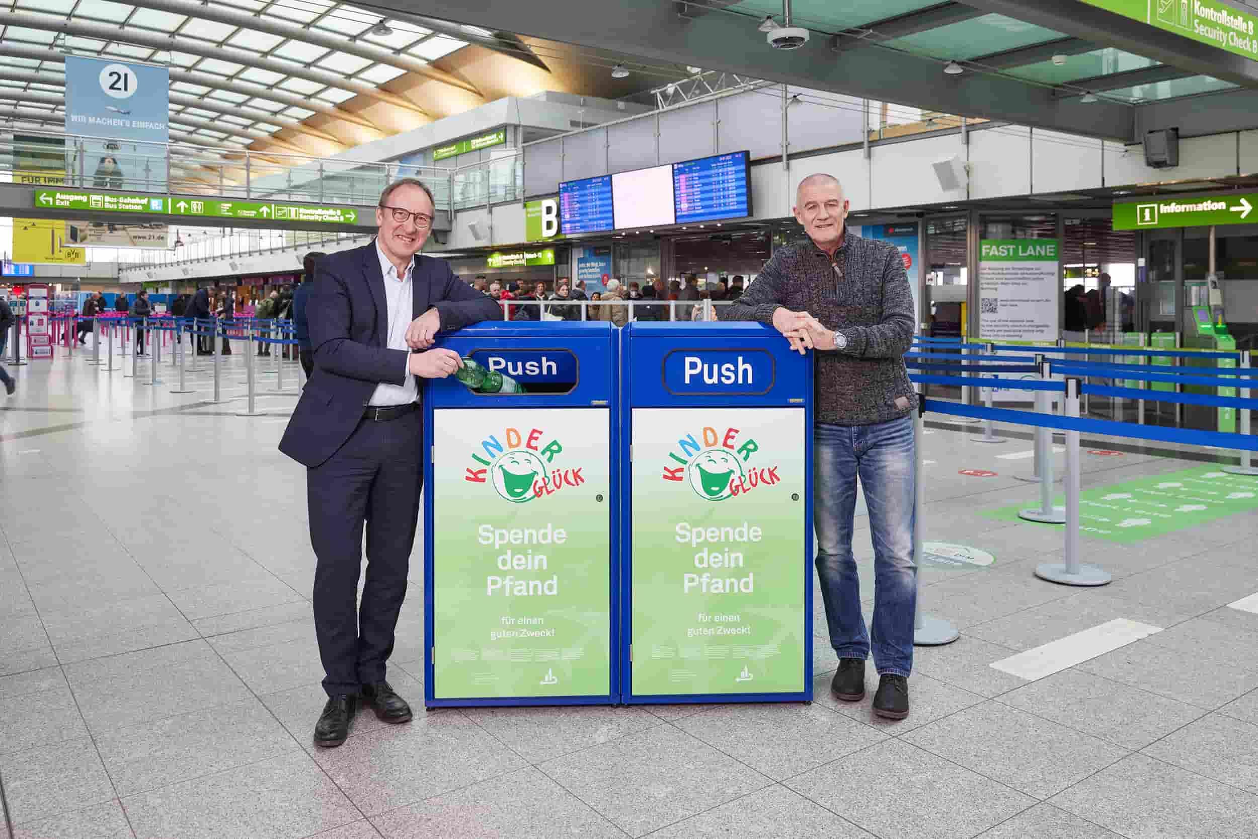 Ludger van Bebber, Geschäftsführer Dortmund Airport und Bernd Krispin, Gründer Stiftung Kinderglück, stehen vor einer Pfandspendenboxbox im Terminal des Dortmund Airport. 