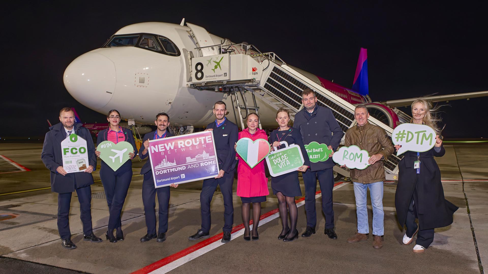 Die Crew des Erstflugs nach Rom sowie Repräsentatnten des Flughafens stehen vor dem Flugzeug und halten Willkommensschilder hoch.