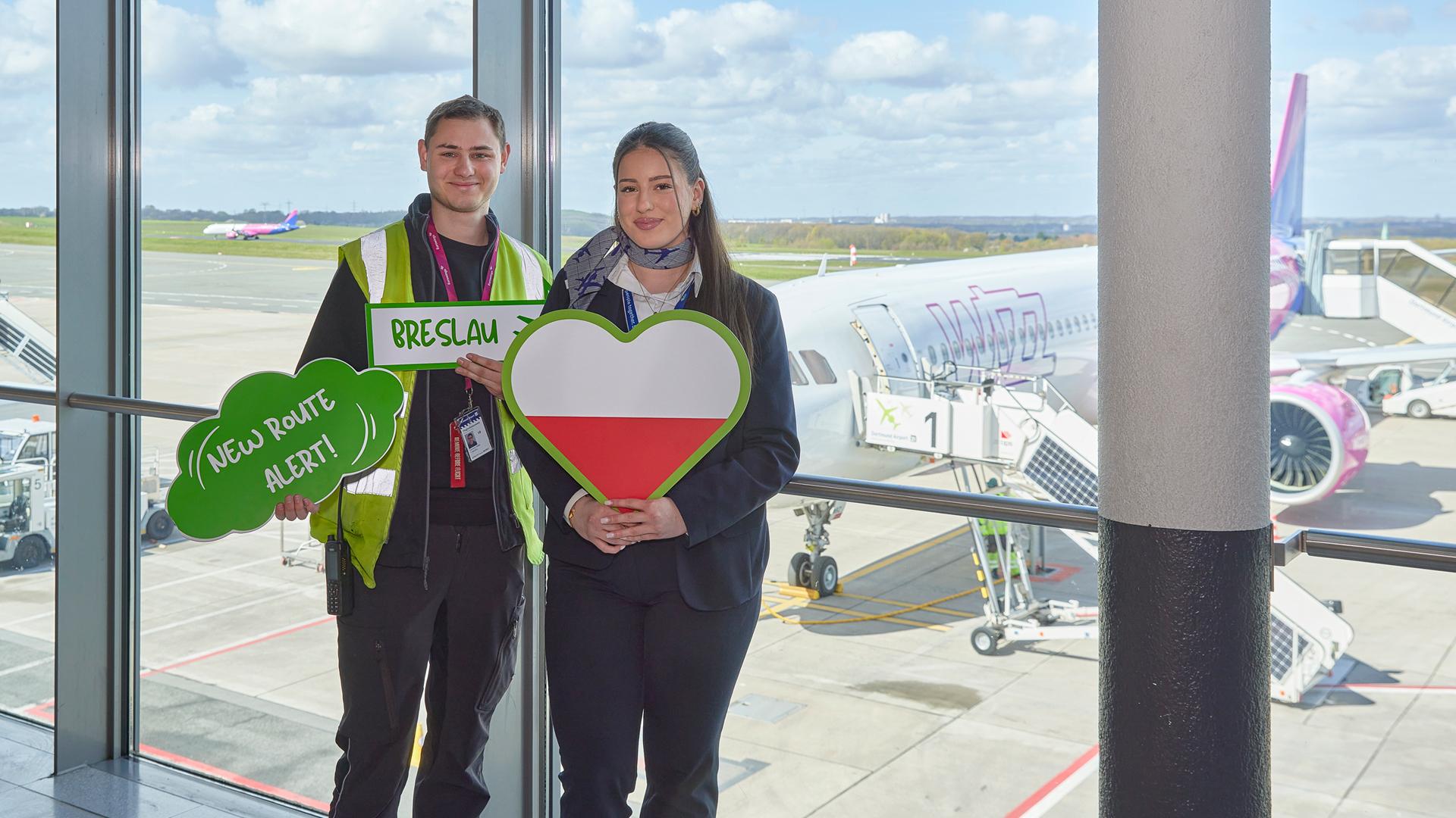 Zwei Mitarbeitende des Dortmunder Flughafens halten Schilder zum Erstflug Breslau hoch. Im Hinterrund ist eine Wizz Air Maschine zu sehen.