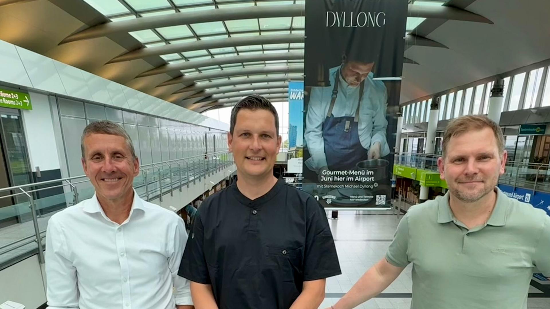 Guido Miletic, Michael Dyllong und Sonnhard Osyka posieren vor einem Banner für das Pop-up-Restaurant von Michael Dyllong am Flughafen