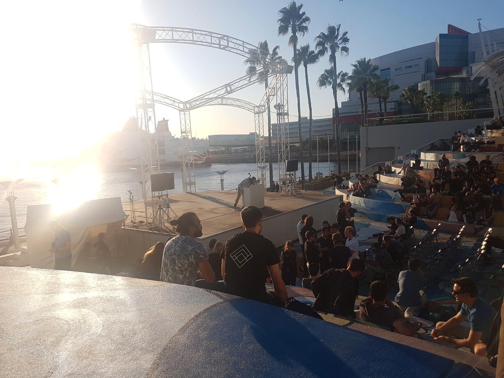 Audience seated beside an outdoor stage at sunset, with palm trees and water behind it.