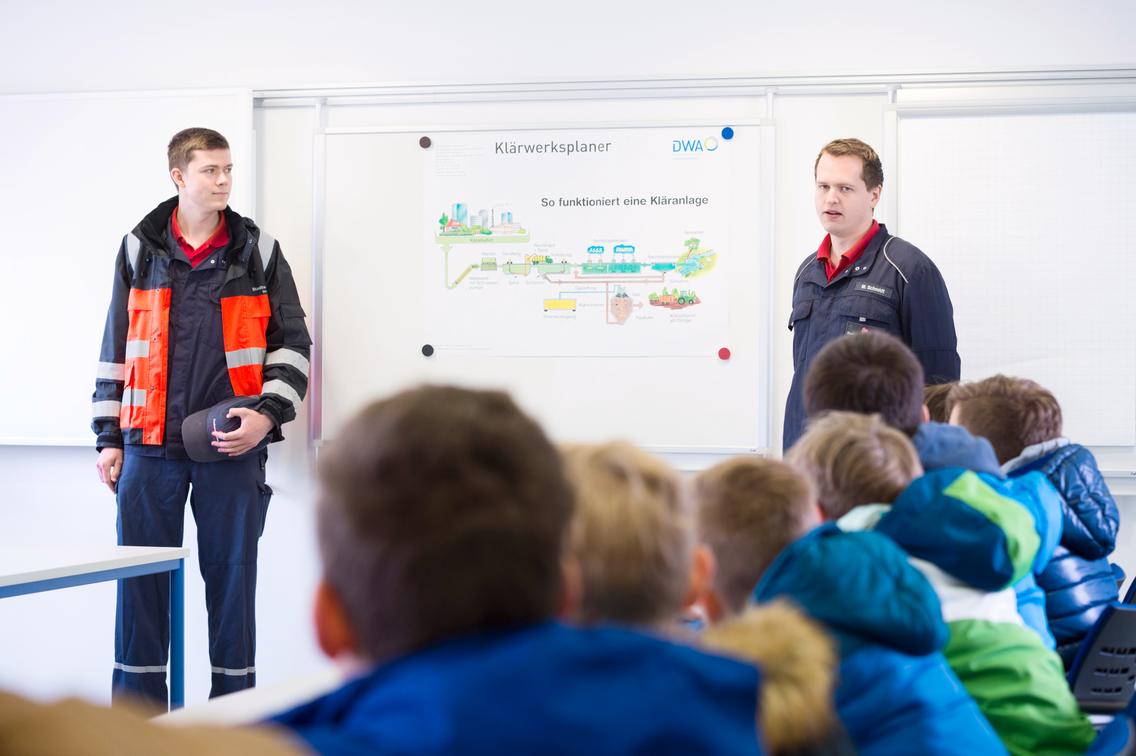 Zwei Männer vor einem Plakat erklären Kindern Klärwerk
