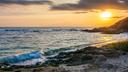 Sonnenuntergang am Goldstrand bei Varna: Sanfte Wellen rollen auf einen ruhigen Sandstrand mit felsigem Ufer, während die tief stehende Sonne den Himmel in warmes Orange und Blau taucht.