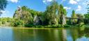 Weitläufige Parklandschaft mit einem ruhigen See im Vordergrund. Auf einem bewachsenen Felsen erhebt sich ein klassizistischer Rundtempel, umgeben von dichten Bäumen und üppigem Grün. Rechts verbindet eine schmale Brücke die Uferbereiche, während sich die Vegetation und die Felsformationen im Wasser spiegeln. Über der Szene spannt sich ein blauer Himmel mit wenigen weißen Wolken.