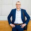 Eine Person in einem blauem Anzug steht mit selbstbewusster Haltung an einer Empfangstheke in einem modernen Firmengebäude. Die Person trägt ein helles Hemd, einen braunen Gürtel und eine Umhängetasche über der Schulter.