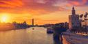 Historischer Turm Torre del Oro am Ufer des Guadalquivir in Sevilla bei Sonnenuntergang. Der Himmel leuchtet in Orange- und Rottönen, Boote fahren über den Fluss, moderne Gebäude und eine Brücke im Hintergrund.