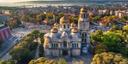 Luftaufnahme der Kathedrale Mariä Himmelfahrt in Varna, Bulgarien: Die markanten goldenen Kuppeln und die detailreiche Architektur stehen im Zentrum, umgeben von Stadtgebäuden, Straßen und einem Hafen mit Kränen im Hintergrund.